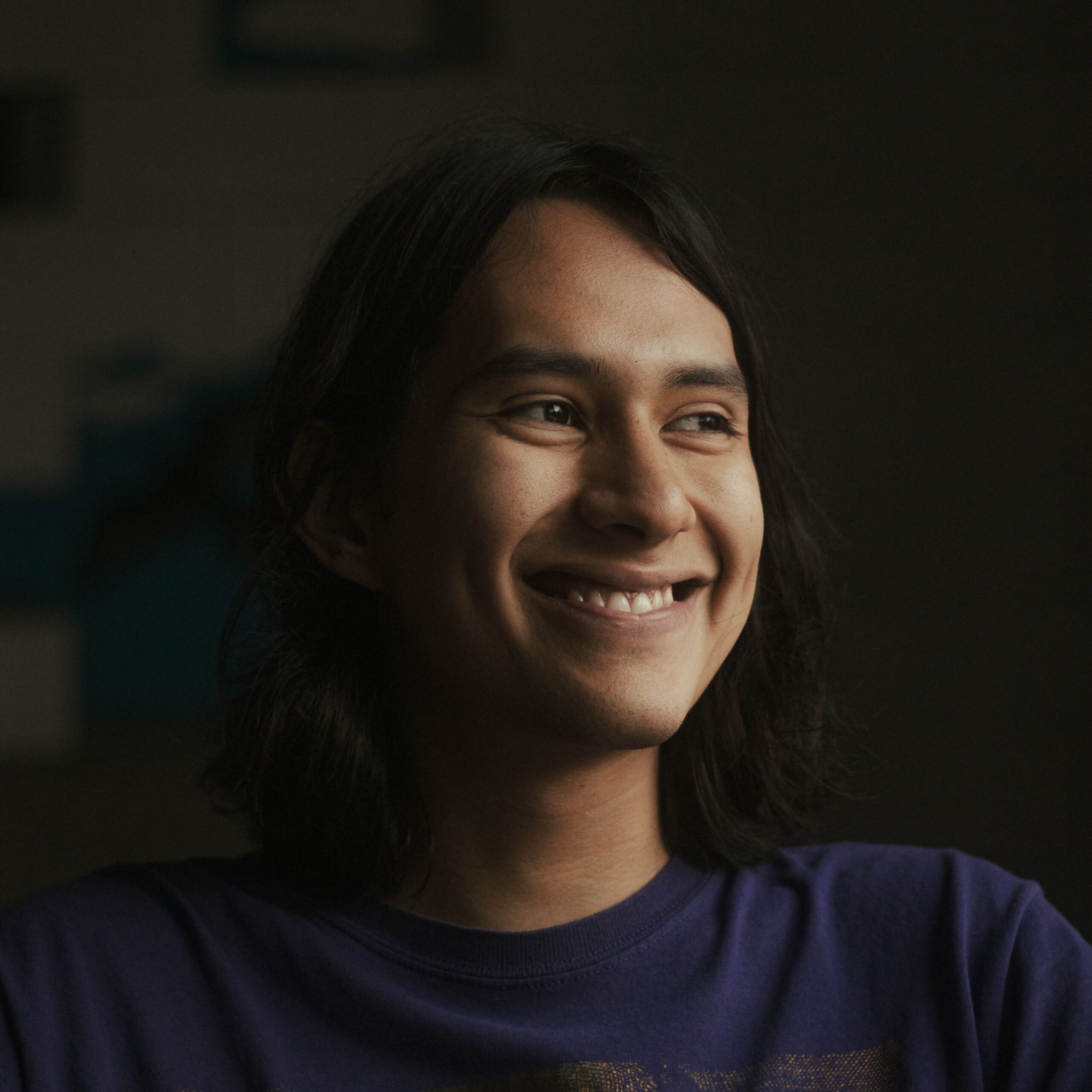 Young man with long hair smiling warmly in a dimly lit indoor setting.